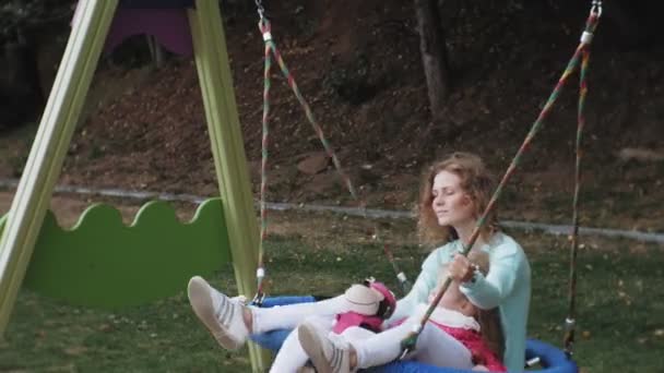 Ei lita jente i rosa kjole og moren hennes som svinger på en rund huske på lekeplassen – stockvideo