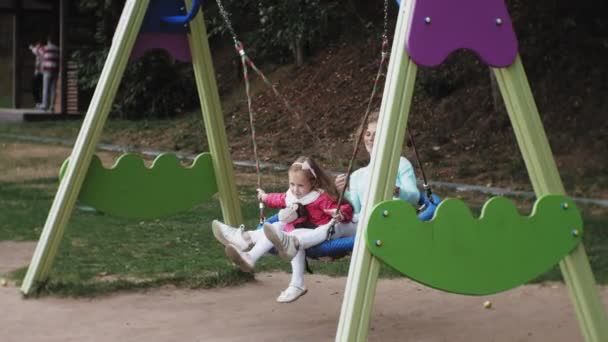 Una niña con un vestido rosa y su madre balanceándose en un columpio redondo en el patio de recreo — Vídeos de Stock