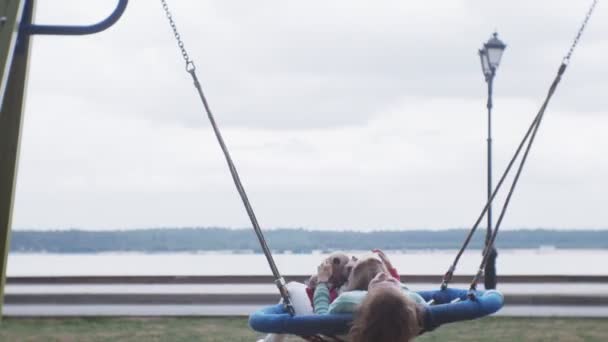 Una bambina con un vestito rosa e sua madre che dondola su un'altalena rotonda al parco giochi — Video Stock