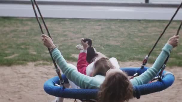 Una bambina con un vestito rosa e sua madre che dondola su un'altalena rotonda al parco giochi — Video Stock