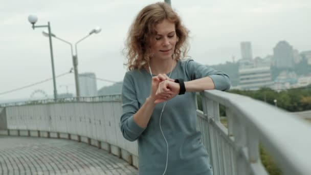 Hübsche lächelnde Fitness-Frau mit Lauf-App vor dem Lauf, um das Tempo auf der Smartwatch zu verfolgen, glücklich lächelndes sportliches Mädchen per SMS vor dem morgendlichen Training — Stockvideo