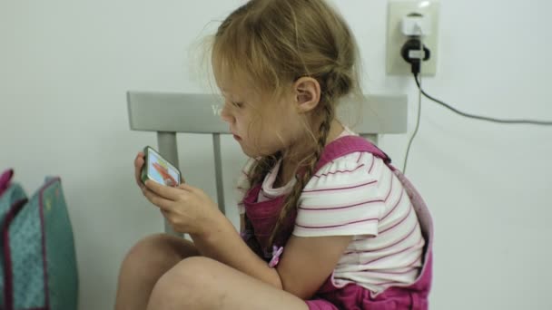 Little girl that sits at the chair, tries to playing a game with her smartphone. Child using mobile phone — Stock Video