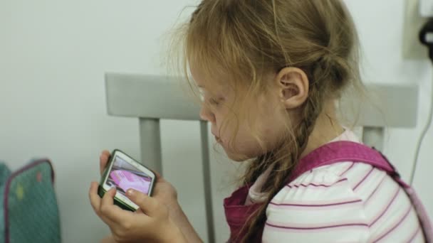 Menina que se senta na cadeira, tenta jogar um jogo com seu smartphone. Criança usando telefone celular — Vídeo de Stock