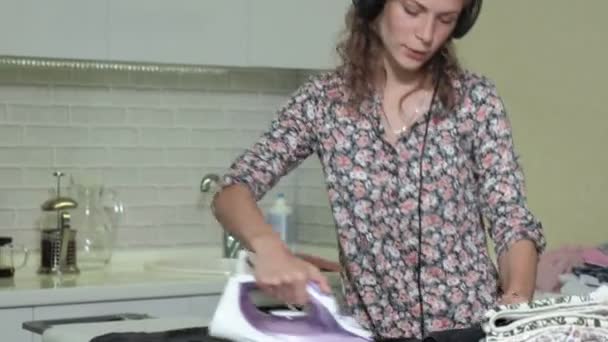Женщина гладит горы прачечной дома на кухне, слушая музыку на наушниках и танцуя — стоковое видео