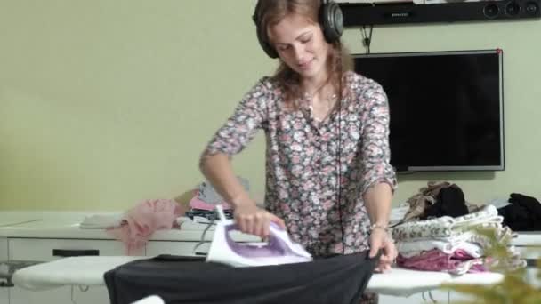 Mujer planchando la montaña de lavandería en casa en la cocina escuchando música en los auriculares y bailando — Vídeos de Stock