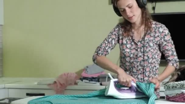 Mujer planchando la montaña de lavandería en casa en la cocina escuchando música en los auriculares y bailando — Vídeo de stock