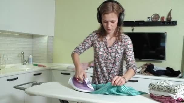 Жінка прасує гору білизни вдома на кухні слухаючи музику на навушниках і танцях — стокове відео