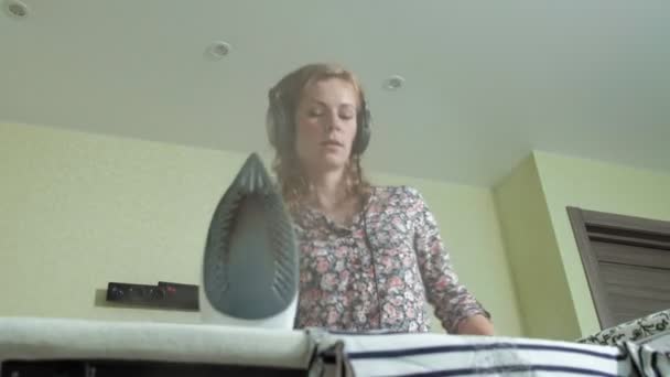 Donna stirare la montagna di lavanderia a casa in cucina ascoltando musica su cuffie e ballando — Video Stock