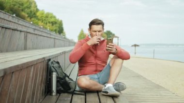 Olgun adam, sahilde ahşap bir bankta oturan bir smartphone kullanarak Turizm