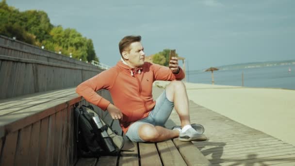 Homme mûr, touriste utilisant un smartphone, assis sur la plage sur un banc en bois — Video