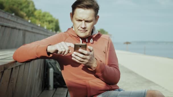 Зрілий чоловік, турист за допомогою смартфона, сидить на пляжі на дерев'яній лавці — стокове відео