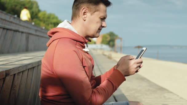 Homem maduro, turista usando um smartphone, sentado na praia em um banco de madeira — Vídeo de Stock