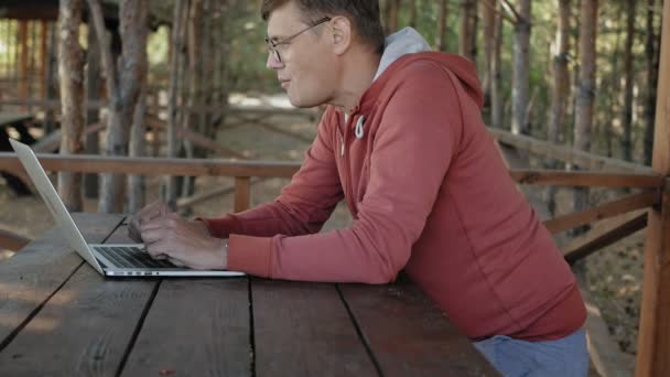 Hombre maduro está trabajando en su computadora portátil al aire libre en la naturaleza durante su vocación forestal envejecido utilizando su computadora portátil para la comprobación de correo electrónico mientras está sentado en la enorme mesa de madera — Vídeos de Stock