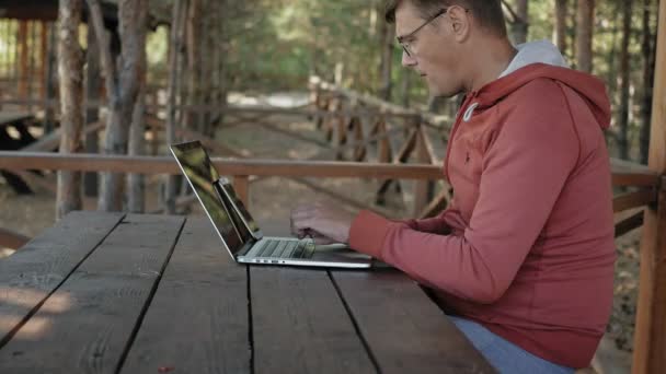 Dojrzały człowiek pracuje na swoim laptopie na zewnątrz w przyrodzie podczas jego powołanie w wieku forester przy użyciu jego laptop dla e-mail sprawdzania siedząc przy wielkim stole drewniane — Wideo stockowe