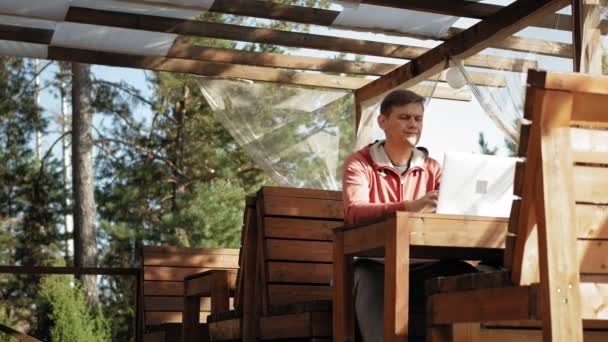 Зрілий чоловік працює на ноутбуці на відкритому повітрі під час свого покликання у віці лісництва, використовуючи свій ноутбук для перевірки електронної пошти, сидячи за величезним дерев'яним столом — стокове відео
