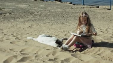 Tatlı bir kız kum üzerine oturur ve bir kitap vuruş bir köpek okur