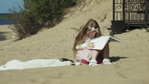 Uma menina doce senta-se na areia e lê um livro acaricia um cão — Vídeo de Stock