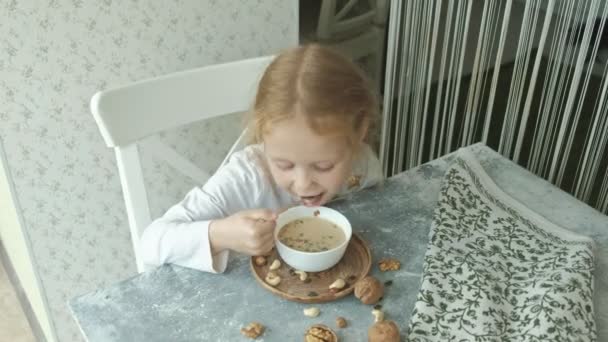 Маленькая милая девочка ест овсянку с орехами и сухофруктами на завтрак. Концепция здорового питания — стоковое видео