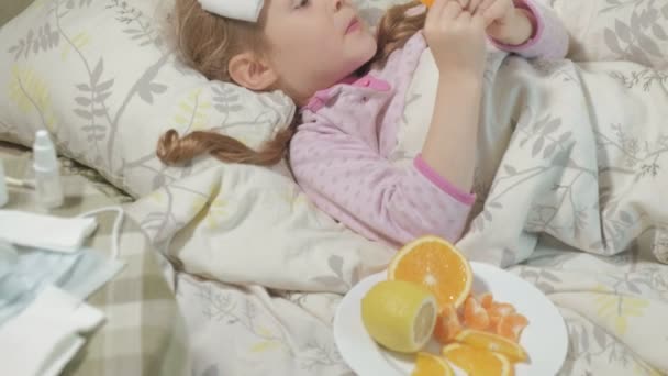 Miúda doente com febre. Uma criança com febre jaz na cama e come frutas . — Vídeo de Stock