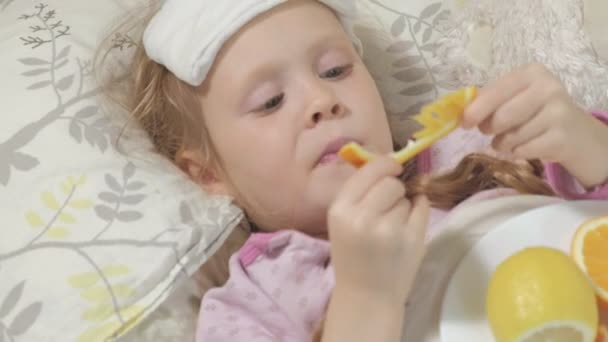 Niña enferma con fiebre. Un niño con fiebre se acuesta en la cama y come fruta. . — Vídeo de stock