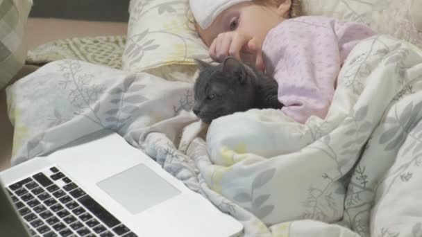 Menina doente com uma temperatura. Uma criança com febre está deitada na cama com um gato . — Vídeo de Stock