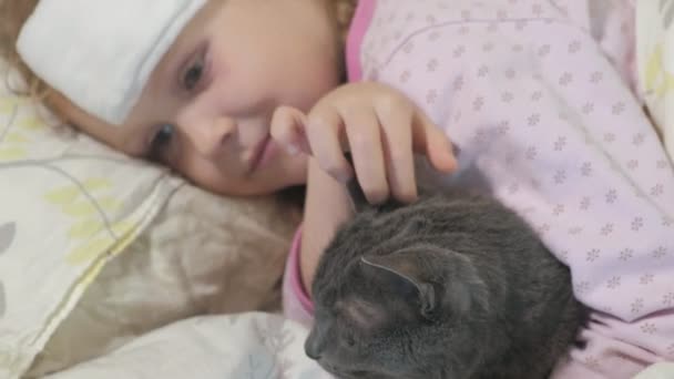 Menina doente com uma temperatura. Uma criança com febre está deitada na cama com um gato . — Vídeo de Stock