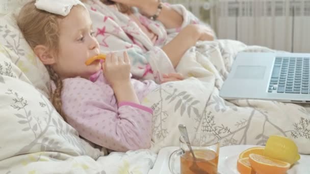 Menina doente com uma temperatura. Criança com febre está deitada na cama com a mãe, comendo frutas e usando um laptop . — Vídeo de Stock