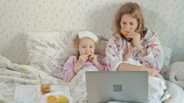 Menina doente com uma temperatura. Criança com febre está deitada na cama com a mãe, comendo frutas e usando um laptop . — Vídeo de Stock