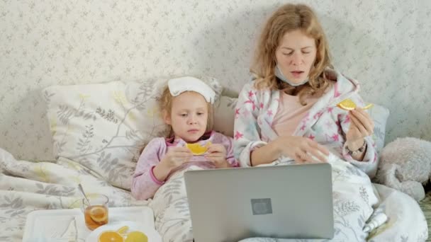 Niña enferma con temperatura. El niño con fiebre está acostado en la cama con su madre, comiendo fruta y usando una computadora portátil . — Vídeo de stock