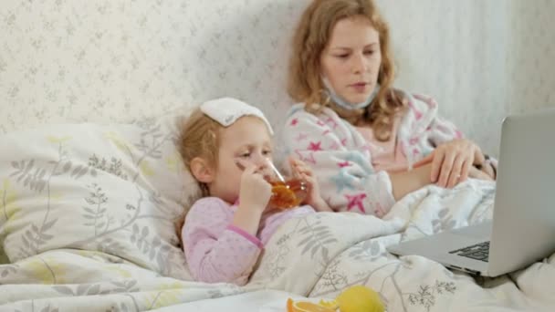 Fille malade avec une température. Enfant avec de la fièvre est couché avec sa mère, manger des fruits et en utilisant un ordinateur portable . — Video