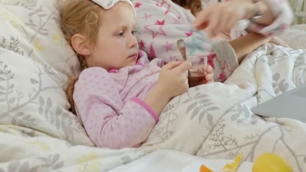 Fille malade avec une température. Enfant avec de la fièvre est couché avec sa mère, manger des fruits et en utilisant un ordinateur portable . — Video