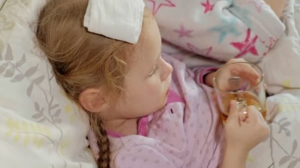 Menina doente com uma temperatura. Criança com febre está deitada na cama com a mãe, comendo frutas e usando um laptop . — Vídeo de Stock