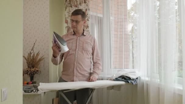 Мужчина гладит свою рубашку по гладильной доске в своем доме — стоковое видео
