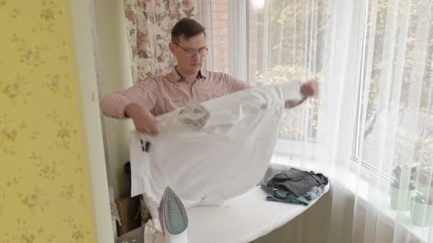 Мужчина гладит свою рубашку по гладильной доске в своем доме — стоковое видео