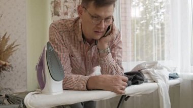 Bir adam onun evinde Ütü masası üzerinde gömleğini konturlar ve bir akıllı telefon 4 k kullanır