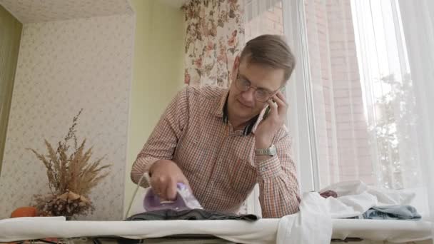 Мужчина гладит рубашку на гладильной доске в своем доме и пользуется смартфоном — стоковое видео