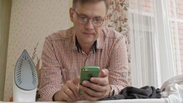 En man stryker skjortan på strykbrädan i sitt hus och använder en smartphone — Stockvideo