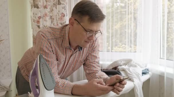 Un uomo accarezza la camicia sull'asse da stiro di casa sua e usa uno smartphone — Video Stock