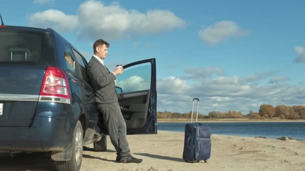 Бизнесмен с чемоданом для ноутбука, отдыхающий на пляже рядом со своей машиной . — стоковое видео