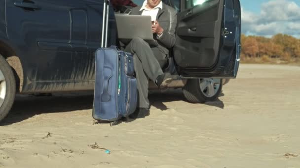 Empresario con una maleta portátil trabajando relajado en la playa cerca de su coche . — Vídeo de stock
