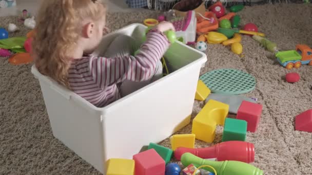 Linda criança rindo, loira pré-escolar, brincando com brinquedos coloridos em uma caixa branca, sentada no chão na sala — Vídeo de Stock