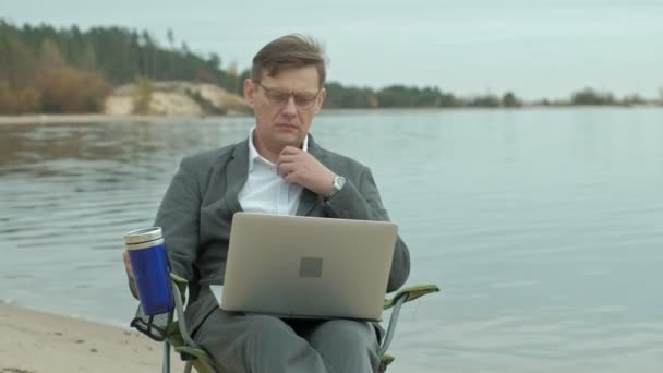 Érett üzletember ül, és pihentető a folyóparton. Férfi öltöny és használ laptop. — Stock videók