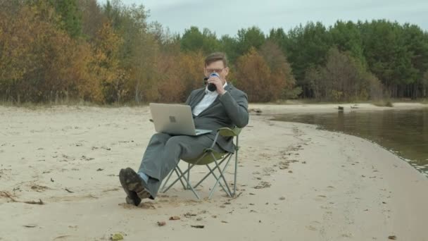 川の近くに座って落ち着いた成熟した実業家。男のスーツとラップトップを使用して. — ストック動画