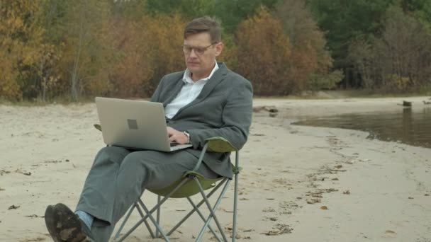 Mature businessman sitting and relaxing near a river. Man in suit and using laptop. — Stock Video