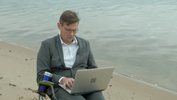 Mature businessman sitting and relaxing near a river. Man in suit and using laptop. — Stock Video