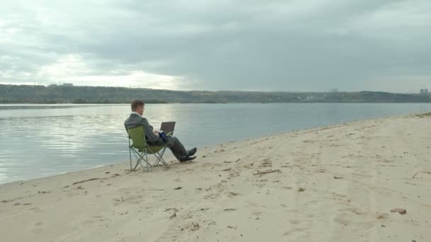 Rijpe zakenman zitten en ontspannen in de buurt van een rivier. Man in pak en met behulp van laptop. — Stockvideo