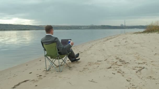 Зрілий бізнесмен сидить і розслабляється біля річки. Людина в костюмі і використовує ноутбук . — стокове відео