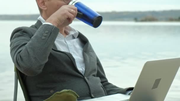 Mogen affärsman sitter och avkopplande nära en flod. Mannen i kostym och med laptop. — Stockvideo