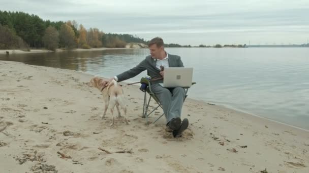 Зрелый бизнесмен с собакой сидит и отдыхает у реки. Человек в костюме и с ноутбуком . — стоковое видео