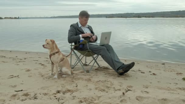 Ώριμες επιχειρηματία με το σκυλί κάθεται και ξεκούραση κοντά σε ένα ποτάμι. Ο άνθρωπος στο κοστούμι και χρησιμοποιούν φορητό υπολογιστή. — Αρχείο Βίντεο